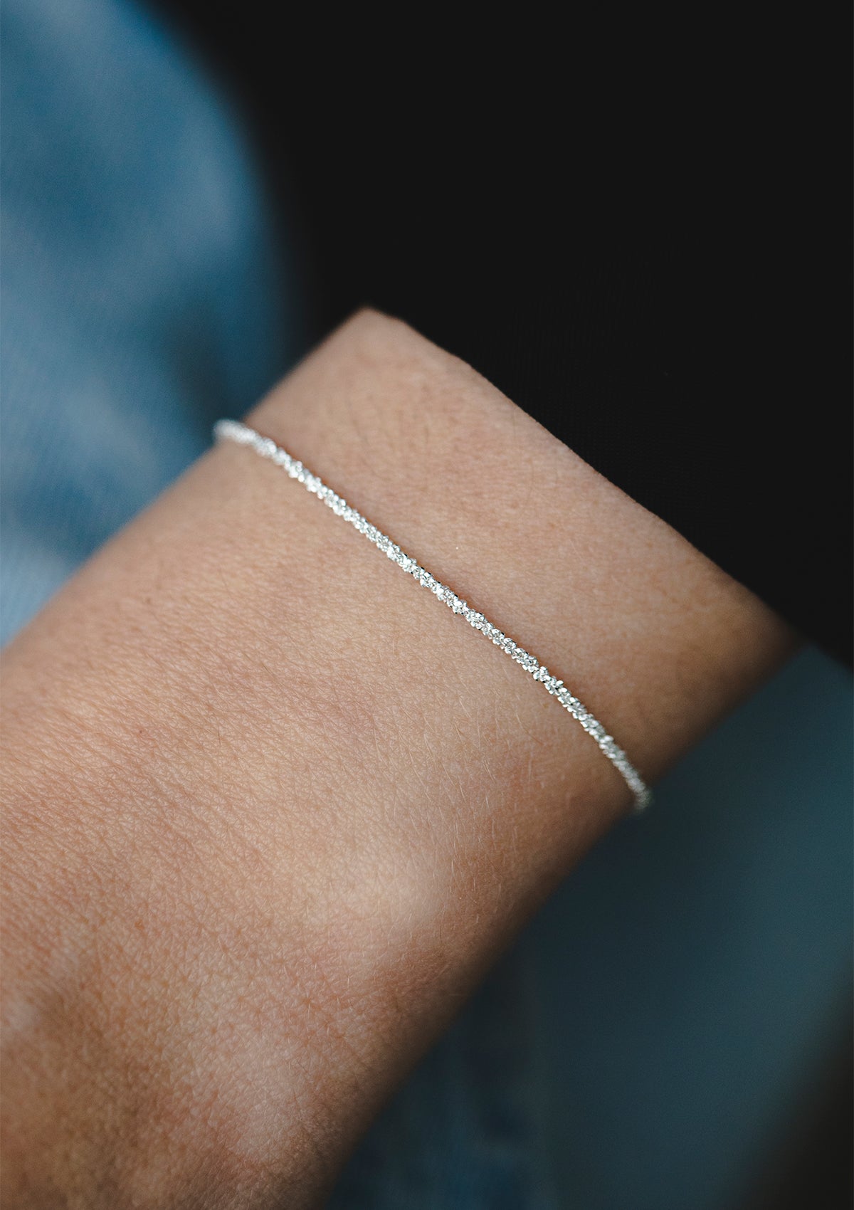 Sparkling sterling silver bracelet on wrist, light-catching fine chain against blue sleeve, refined and simple look