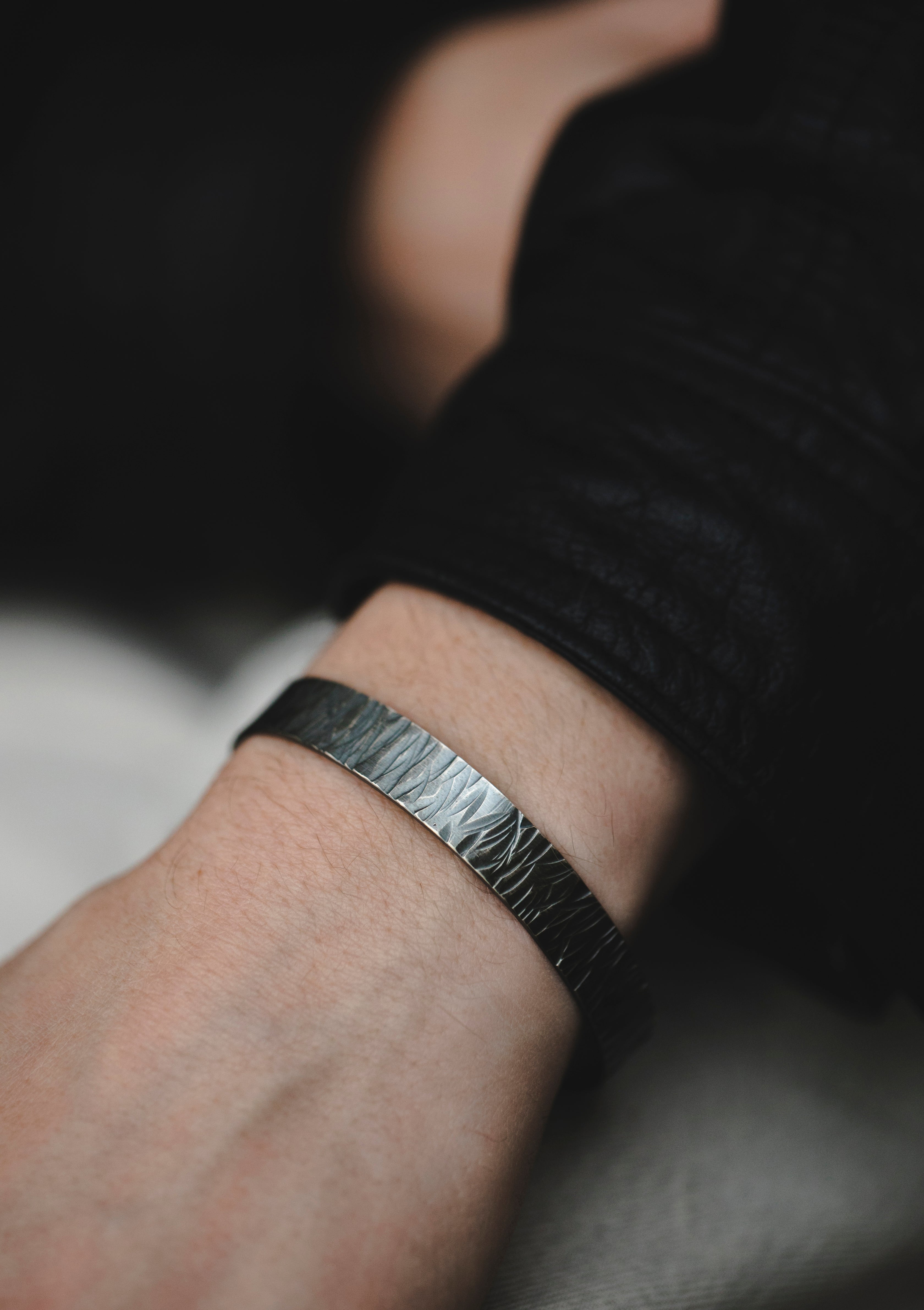 Men’s hammered silver cuff bracelet worn on wrist, blackened matte finish, styled with casual knitwear