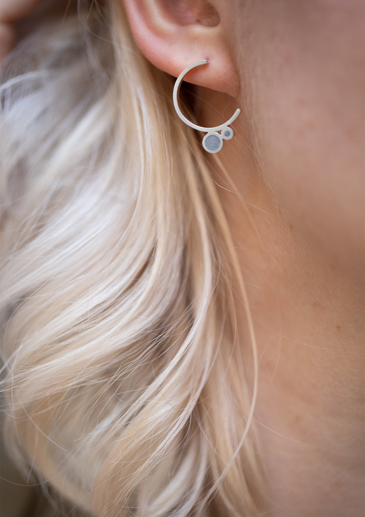 Contemporary silver hoop earring with concrete inlay, worn by blonde model with waves, close-up side profile, minimalist design focus