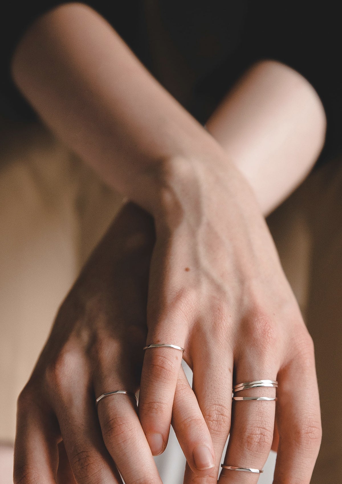 Close-up of crossed hands adorned with multiple thin silver stacking rings, soft light highlights the delicate handcrafted texture and geometric lines.