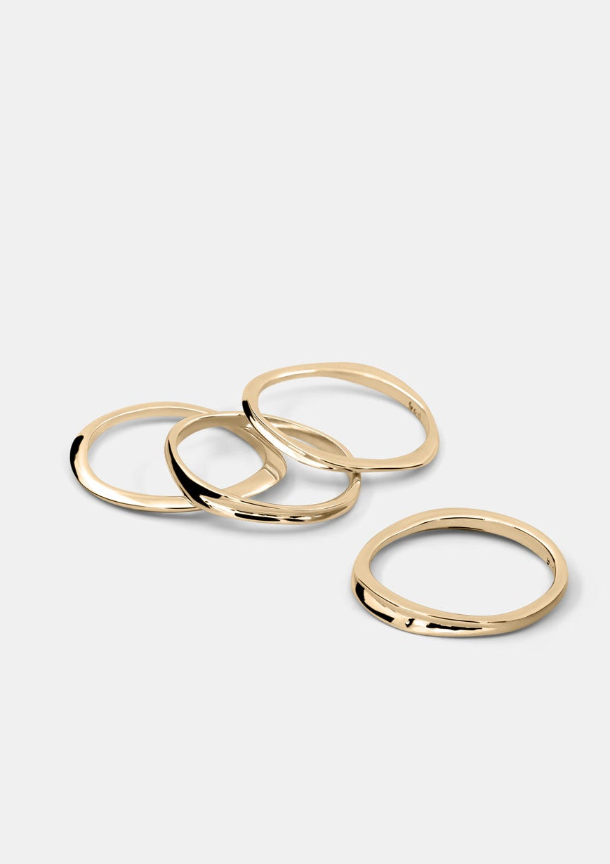 Four polished gold-toned metal rings of varying oval and circular sizes, arranged on a light gray background.